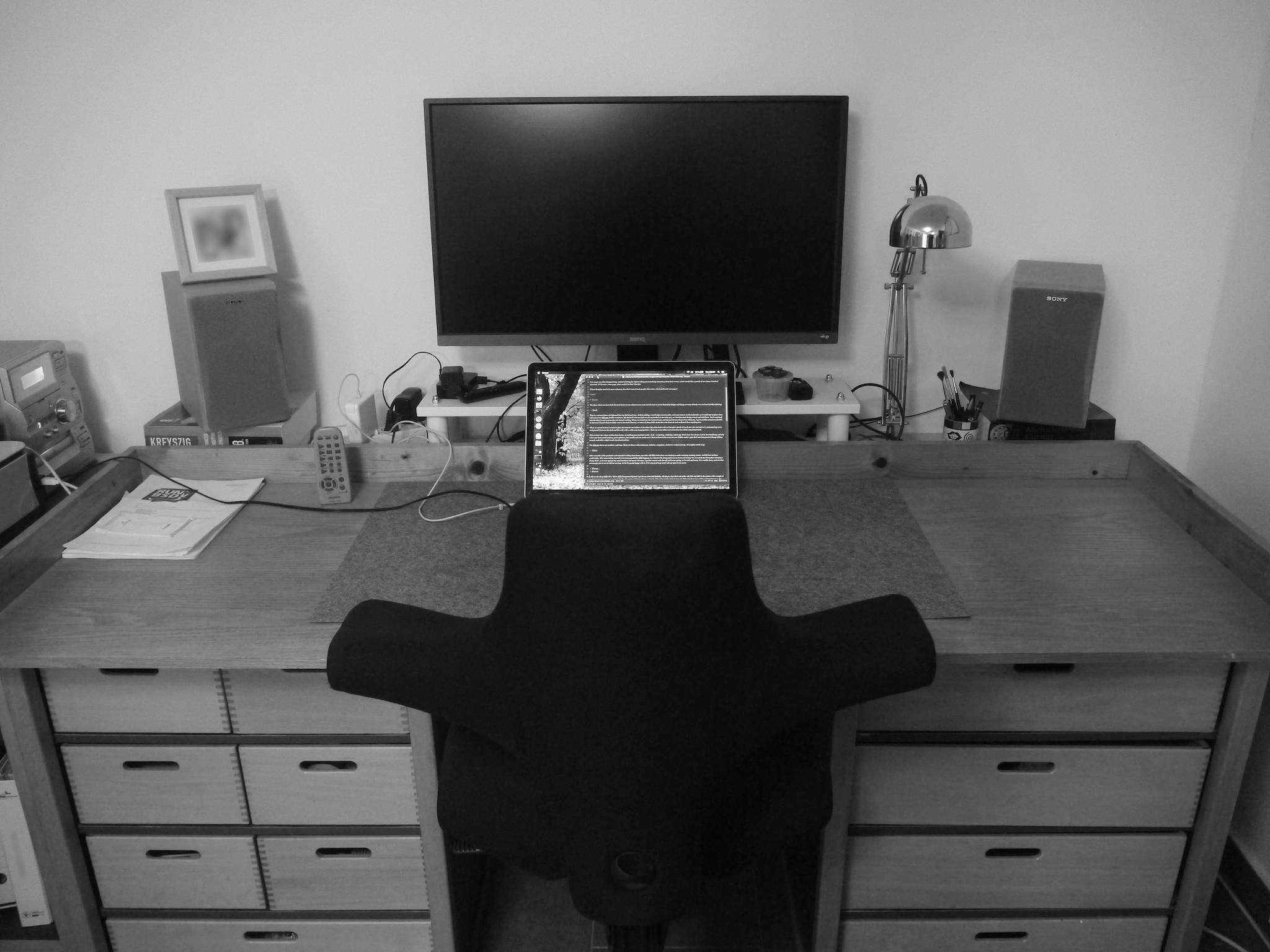 Black and white photo of a simple wooden desk on two sets of draws. In the middle sits a laptop, and behind it a large black screen. Left and right are stereo speakers. Black and white photo of a simple wooden desk on two sets of draws. In the middle sits a laptop, and behind it a large black screen. Left and right are stereo speakers.